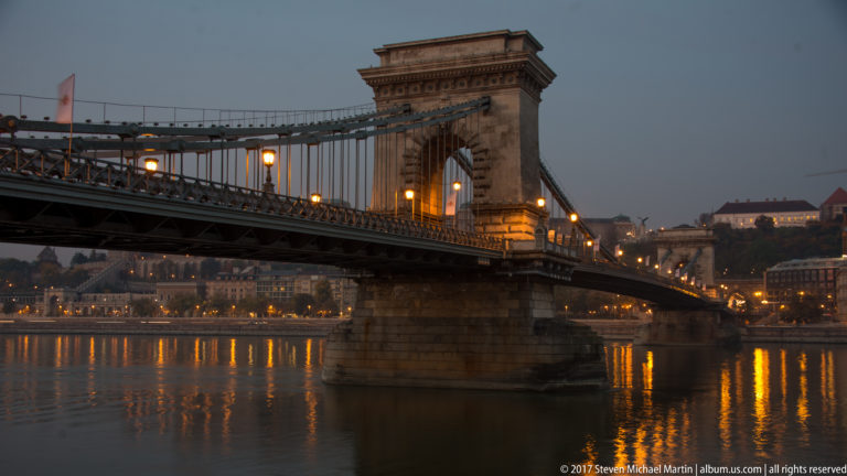 Blue-hour view of Chain Bridge with illuminated stone tower, suspension chains, and reflections in the Danube.