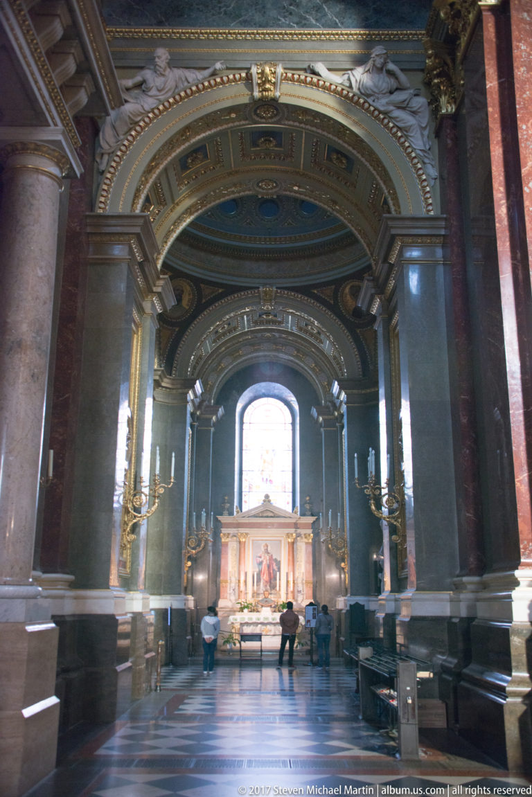 Long basilica side aisle with red marble columns, arches, candelabra, and a lit altar at the far end.