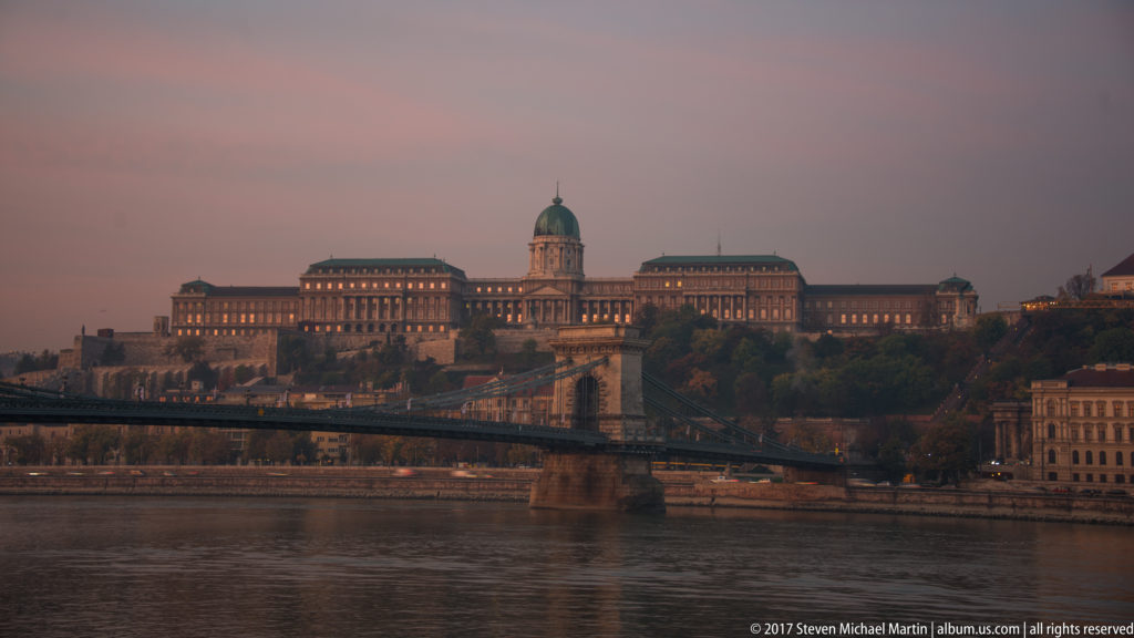 Dusk view of Buda Castle above the Danube with Chain Bridge crossing the foreground.