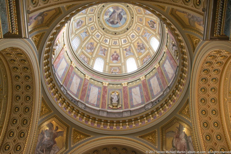 Vertical view of St. Stephen’s Basilica dome with paintings, windows, gilded bands, and figurative pendentives.