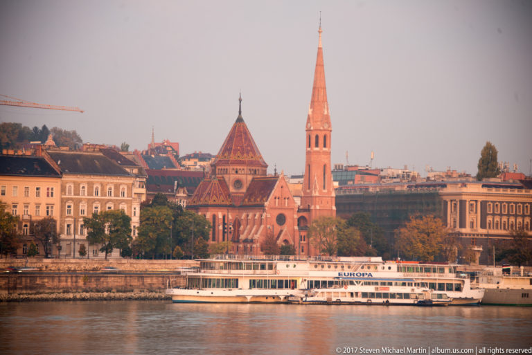 Red-brick Reformed Church with patterned roof and tall slender spire beside the Danube, with a white riverboat in front.