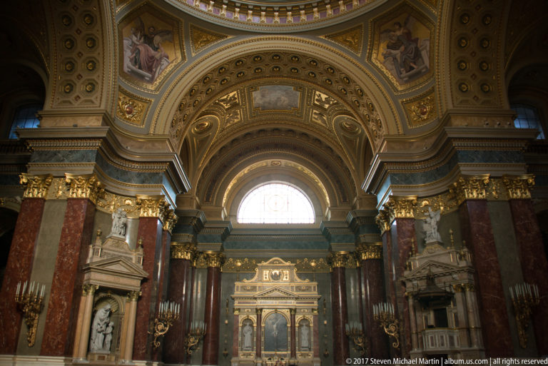 Centered basilica interior with red marble piers, gilded capitals, arched window, side monuments, and altar in the distance.