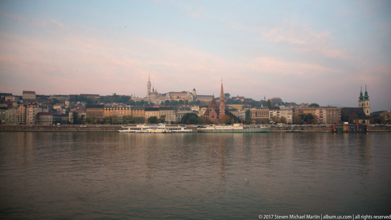 Wide evening view across the Danube toward Buda’s historic riverfront, church spires, hillside buildings, and a white riverboat.