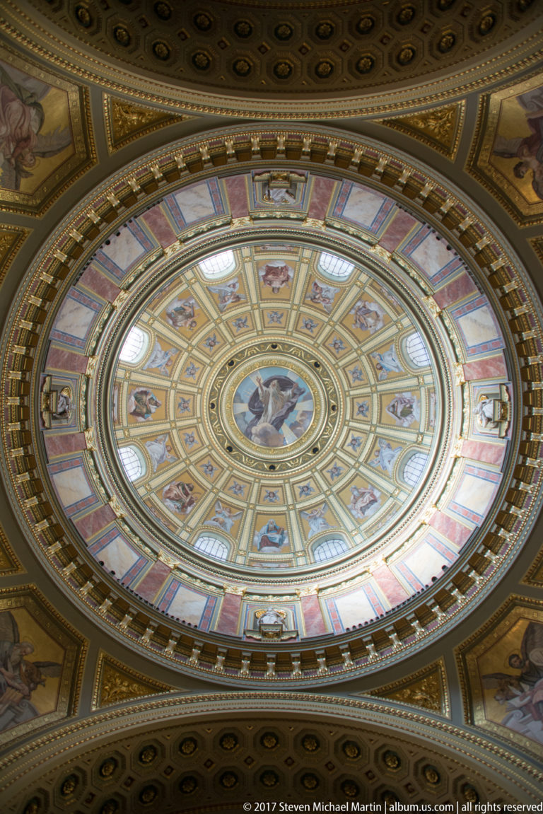 Direct upward view into St. Stephen’s Basilica dome with painted figures, gilded coffers, windows, and central medallion.