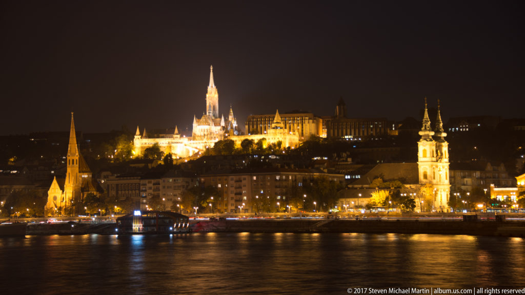 Night panorama across the Danube toward illuminated Castle Hill, Matthias Church, and twin church towers on the Buda riverfront.