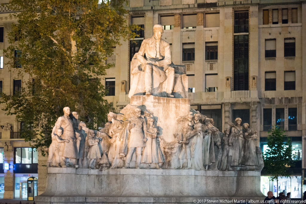 Large pale stone monument with a seated figure above many standing and kneeling figures in an urban square.