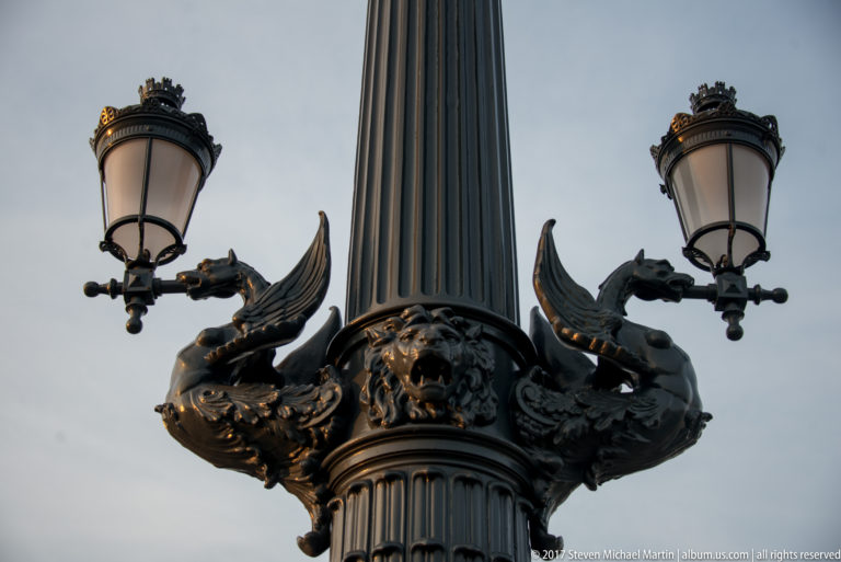 Low-angle ornate bridge lamppost with two lanterns and paired dragon-like cast-metal creatures around a fluted column.