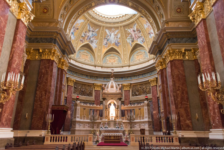 Main sanctuary of St. Stephen’s Basilica with red marble piers, painted dome, gilded capitals, and illuminated altar.