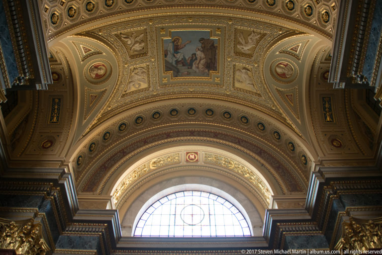 Painted and gilded church vault above a large semicircular window with figurative panels and decorative coffers.