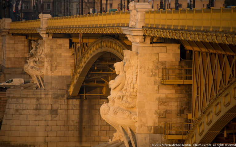 Close detail of Margaret Bridge showing carved stone figures, masonry pier, and iron arch structure in warm light.