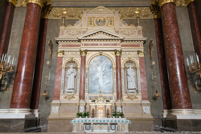 Marble side altar with two standing saint statues, red columns, gilded ornament, candles, and a central devotional panel.