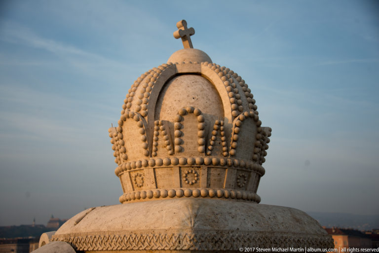 Close stone carving of the Hungarian Holy Crown with beaded arches and a small cross against blue sky.