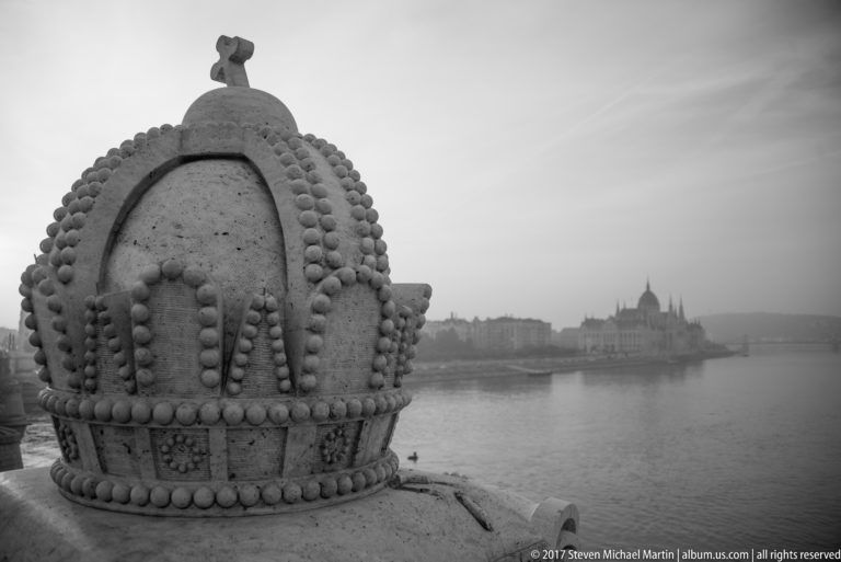 Stone carving of the Hungarian Holy Crown in the foreground with the Parliament Building hazy across the Danube.