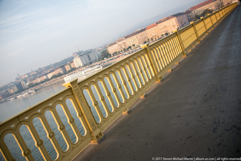 Diagonal view along an ornate yellow-painted bridge railing with the Danube and city buildings beyond.