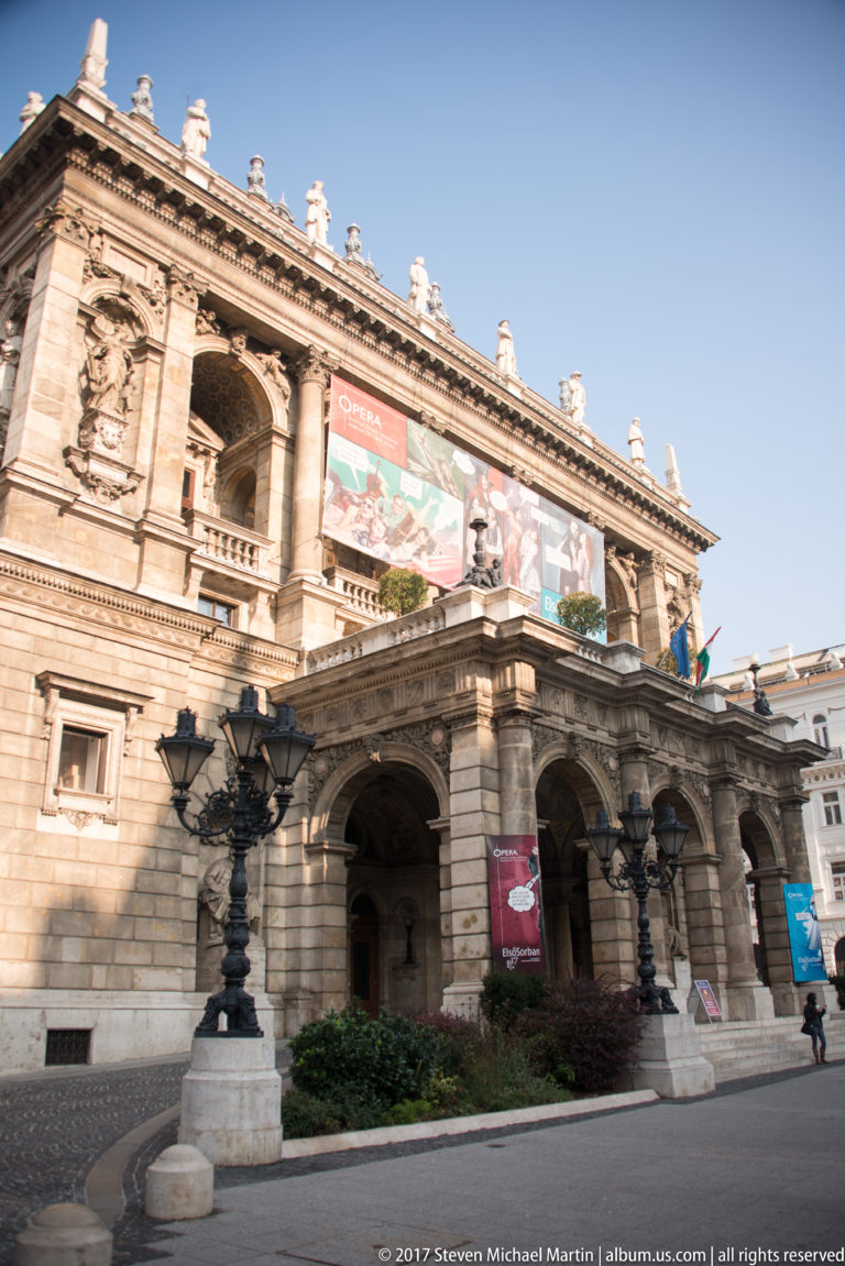 Street-level Hungarian State Opera House façade with arched entrance, ornate lamps, stone sculpture, and performance banners.