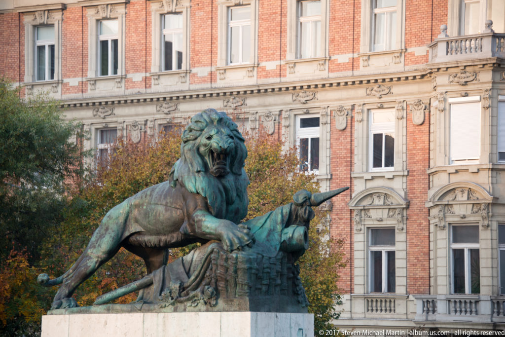 Patinated bronze lion standing over a recumbent sculptural form in front of an ornate red-brick building.