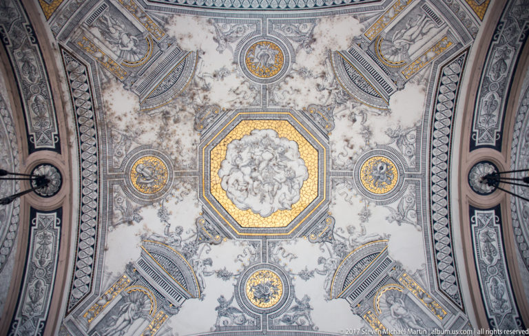 Symmetrical ornate ceiling with central octagonal figurative painting, gilded medallions, gray-blue scrollwork, and light fixtures.