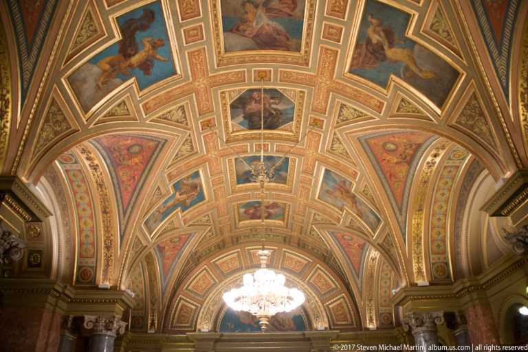 Long gilded and painted vaulted ceiling with figurative panels, medallions, floral ornament, and chandelier.