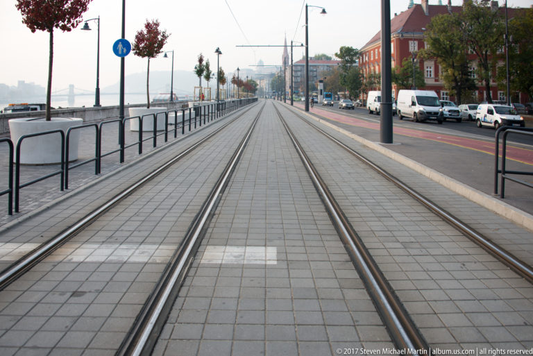 Straight tram tracks and overhead wires along the Buda Danube embankment, with river to the left and road traffic to the right.
