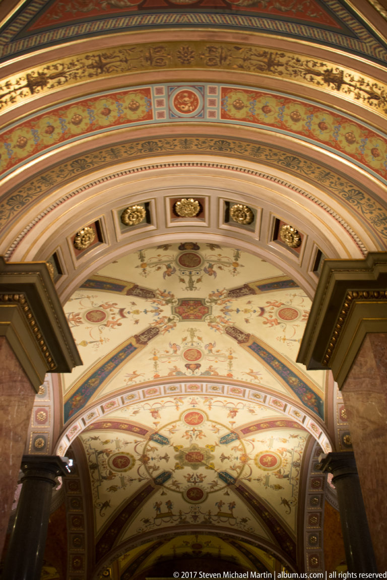 Painted and gilded opera-house ceiling with floral panels, rosettes, arches, and classical ornament.