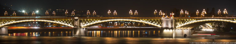 Night view of Margaret Bridge with illuminated iron arches and golden reflections across the Danube.