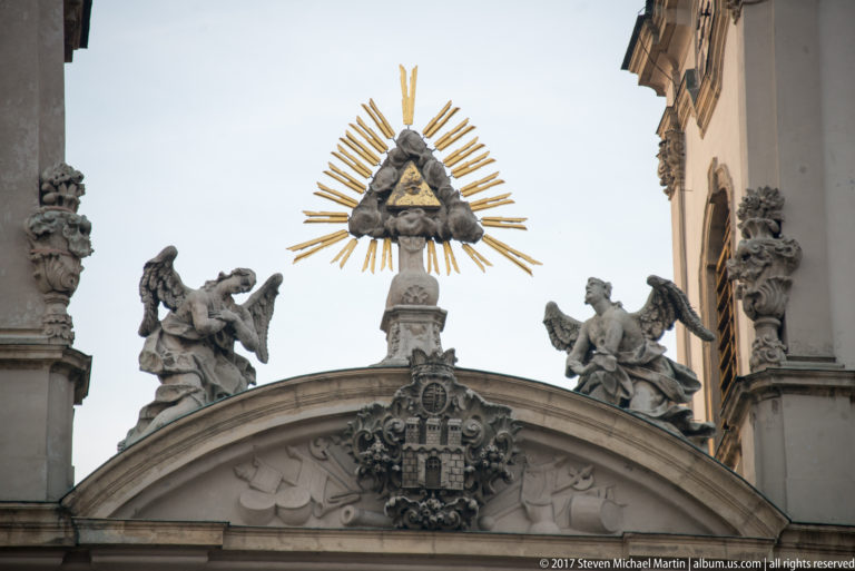Rooftop pediment with winged stone figures, central coat of arms, and a radiant triangular emblem above.