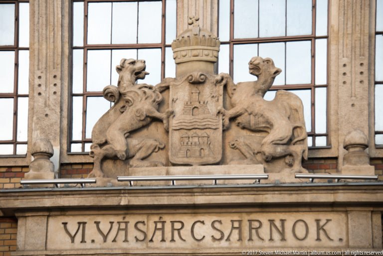 Stone façade detail with the word VÁSÁRCSARNOK beneath a coat of arms supported by two carved animals.