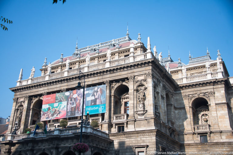 Stone façade of the Hungarian State Opera House with arcades, statues, balustrades, and colorful performance banners.