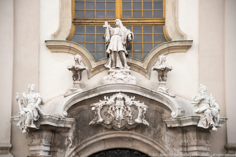 Ornate Baroque doorway with white stone religious figures, heraldic decoration, scrollwork, and a large window above.