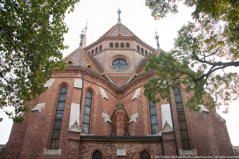 Red-brick church rear elevation with tall windows, patterned tiled roofs, circular window, and decorative roof finials.