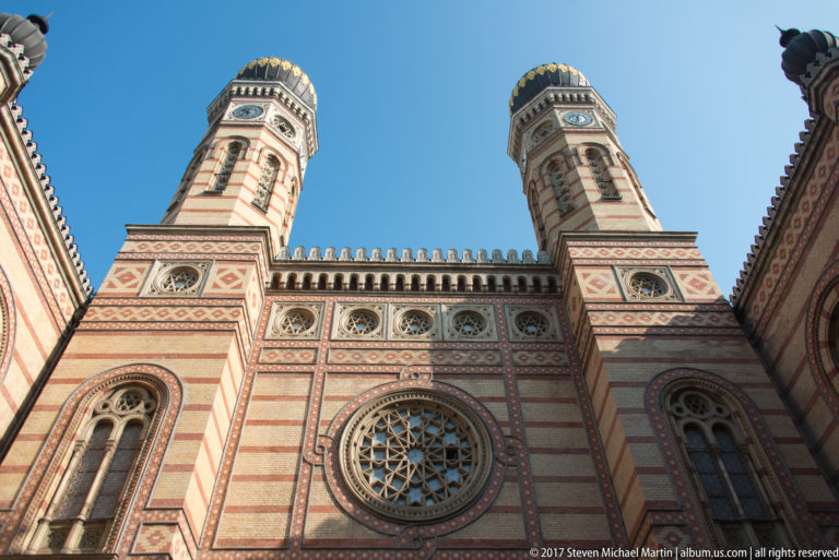 Low-angle view of the Great Synagogue with twin towers, striped brickwork, circular windows, and geometric ornament.