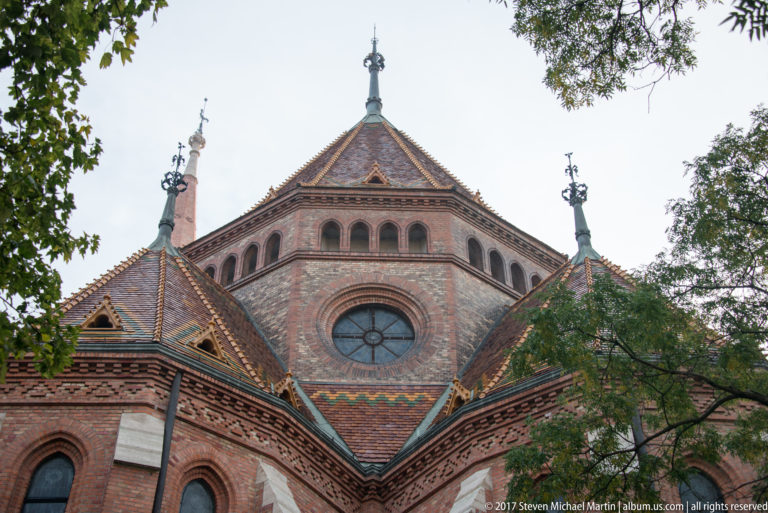 Red-brick church with circular and arched windows, patterned tile roofs, metal finials, and leafy trees.