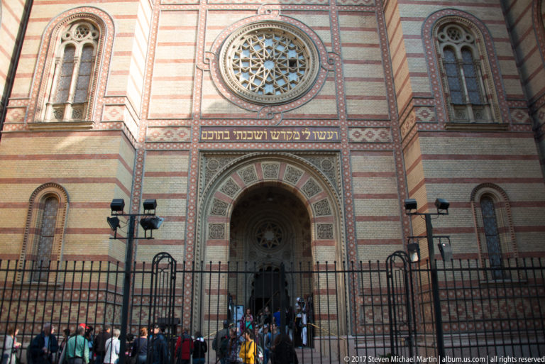 Visitors at the Dohány Street Synagogue entrance beneath Hebrew lettering, striped masonry, and a circular ornamental window.