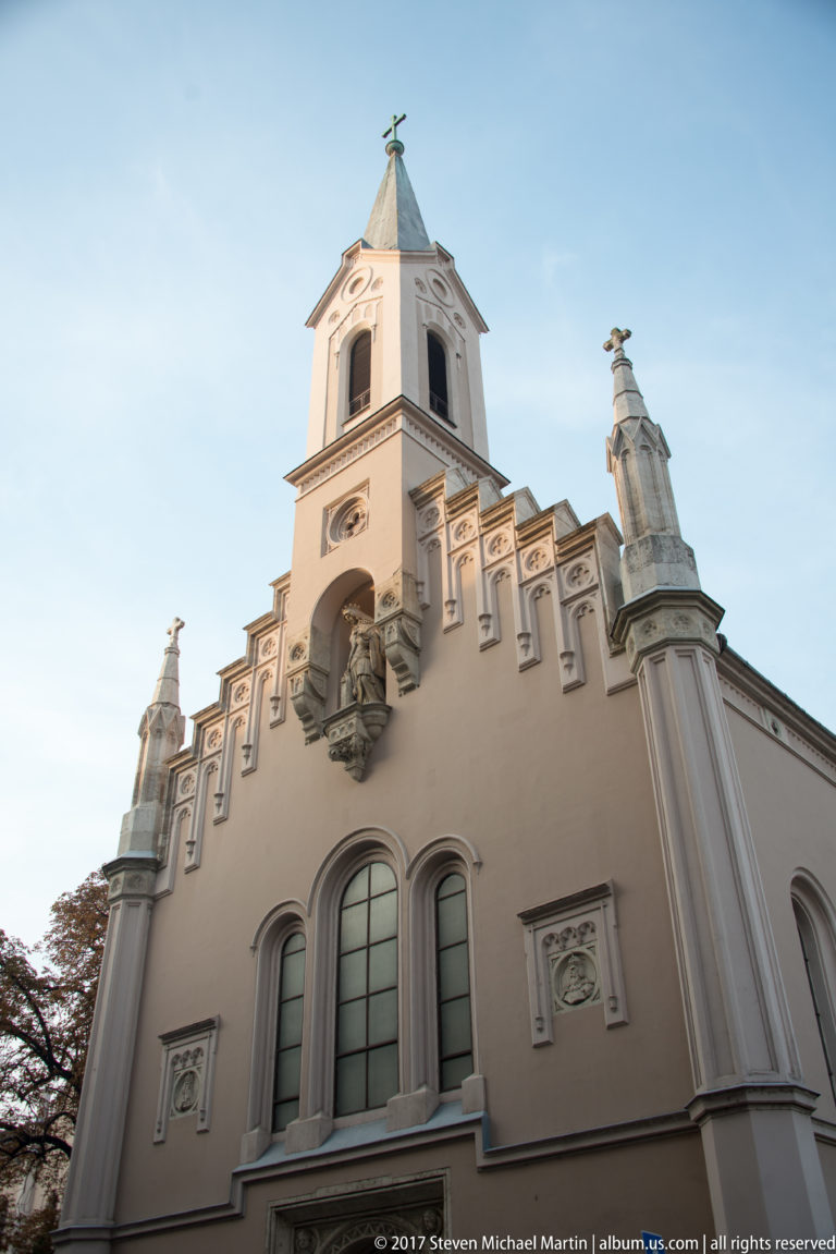 Low-angle pale church façade with square tower, arched windows, rooftop statues, and decorative gable.