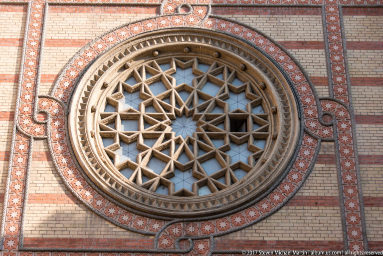 Large circular synagogue window with intricate geometric tracery surrounded by patterned brick and stone bands.
