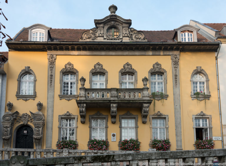 Symmetrical yellow historic townhouse with ornate stone windows, central balcony, carved doorway, and flower boxes.