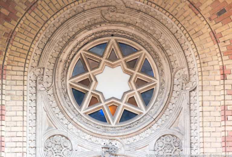 Star of David set within a circular synagogue window surrounded by carved stone ornament and brickwork.