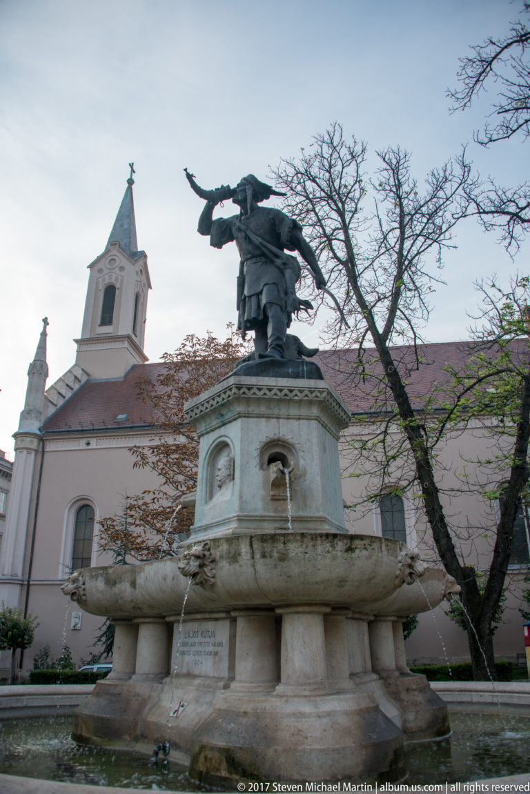 Bronze winged or draped figure atop a circular stone fountain with a pale church and autumn trees behind.
