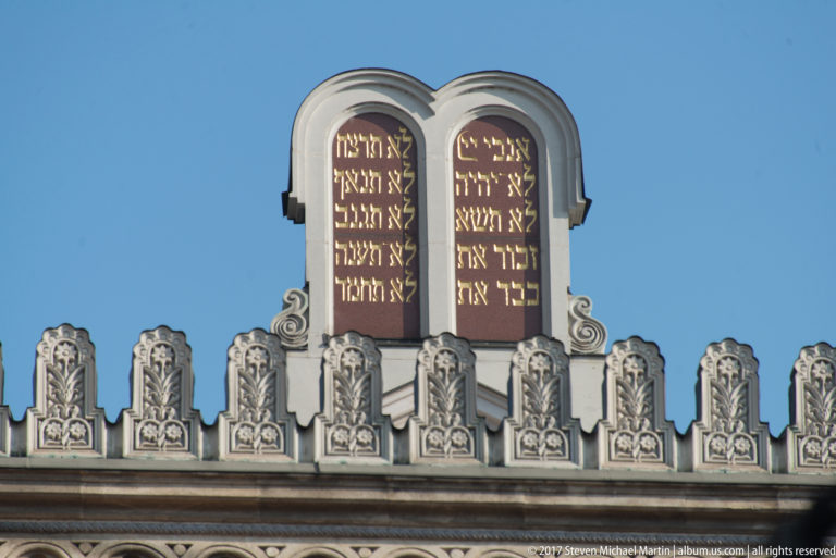 Hebrew-inscribed tablets of the Ten Commandments above an ornate synagogue roofline against blue sky.