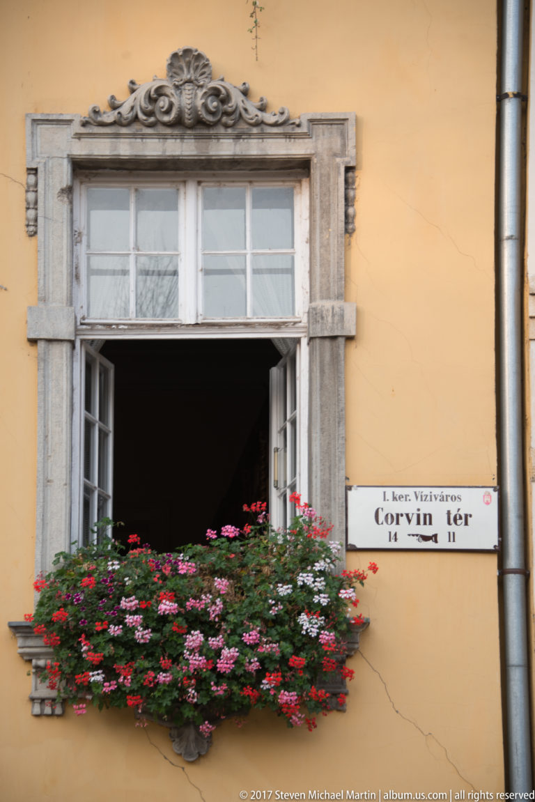 Open stone-framed window with red and pink geraniums beside a street sign reading Corvin tér.