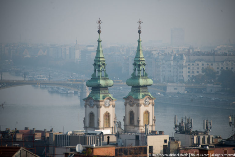 Twin green Baroque towers of Saint Anne Church with the Danube and Pest cityscape behind them.