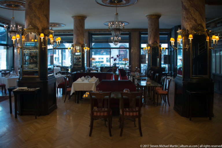 Formal restaurant interior with dark wood paneling, marbleized columns, chandeliers, parquet floor, mirrors, and white tablecloths.