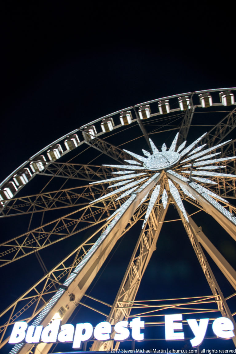 Low-angle night view of Budapest Eye showing bright sign, steel supports, radiating spokes, and lit cabins.