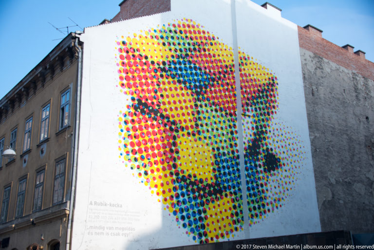 Large colorful dotted portrait mural on a white wall with faint Hungarian text referencing the Rubik’s Cube.
