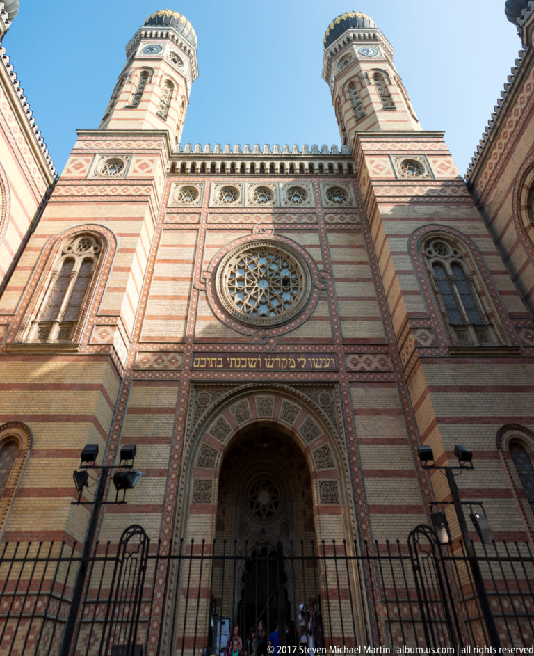 Low-angle façade of Dohány Street Synagogue with striped brickwork, large rose window, Hebrew inscription, and twin domed towers.