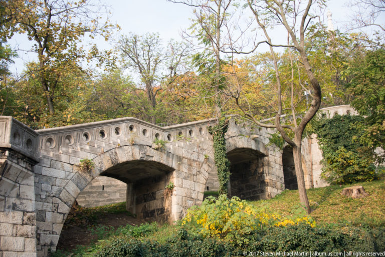 Small two-arched stone footbridge with circular parapet openings surrounded by autumn trees and garden plants.