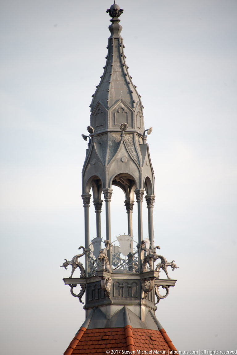 Ornate rooftop turret with slender columns, decorative metalwork, finials, and a steep canopy above red roof tiles.