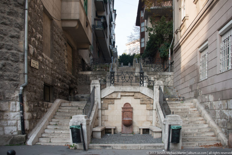 Stone stairway rising between Buda buildings around a small central wall fountain and wrought-iron railings.