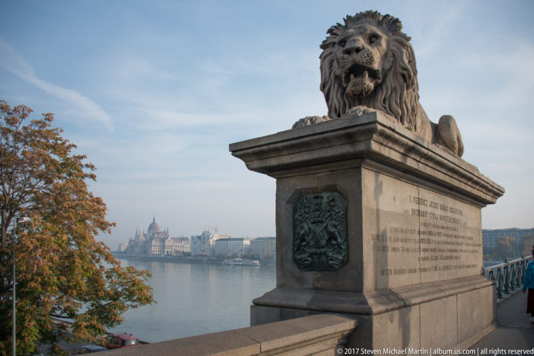 Stone Chain Bridge lion on a pedestal with the Danube and Hungarian Parliament Building in the distance.