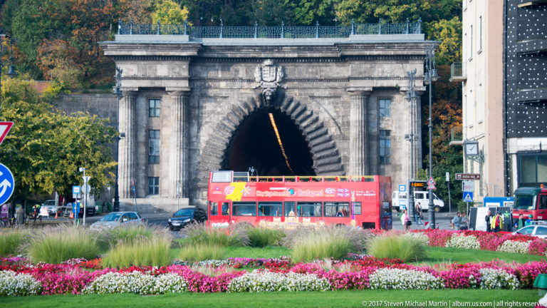 Stone arched portal of the Castle Hill Tunnel with a red sightseeing bus and flower beds in front.